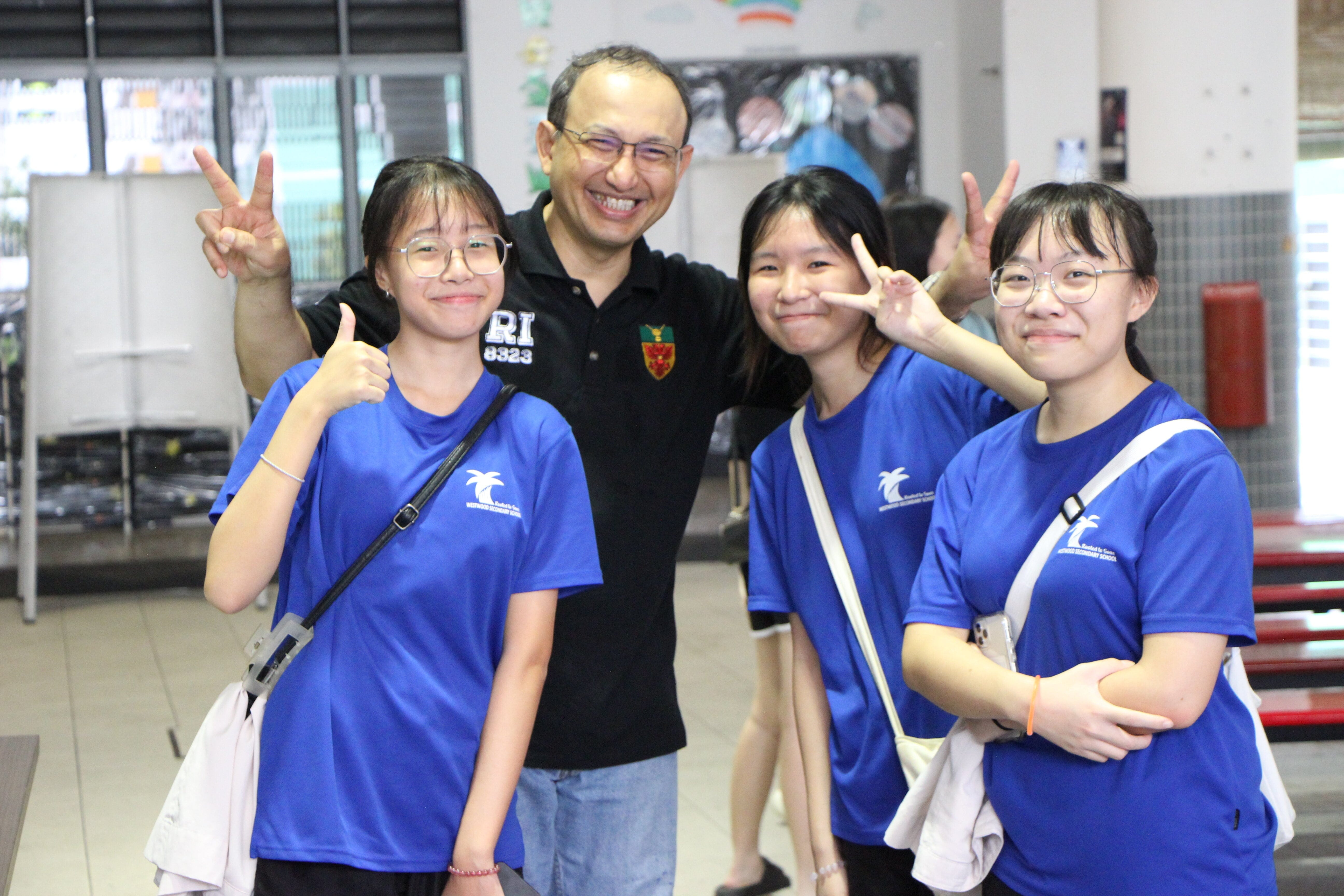 Three girls in blue school shirts and a smiling man in black pose with peace signs indoors.